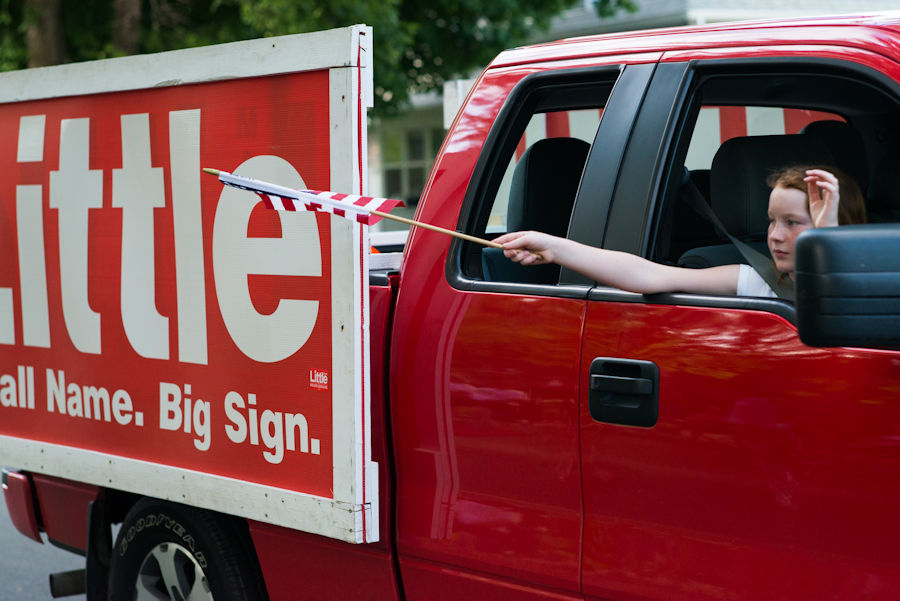 Yard sign on the side of a truck with girl waving flag in window
